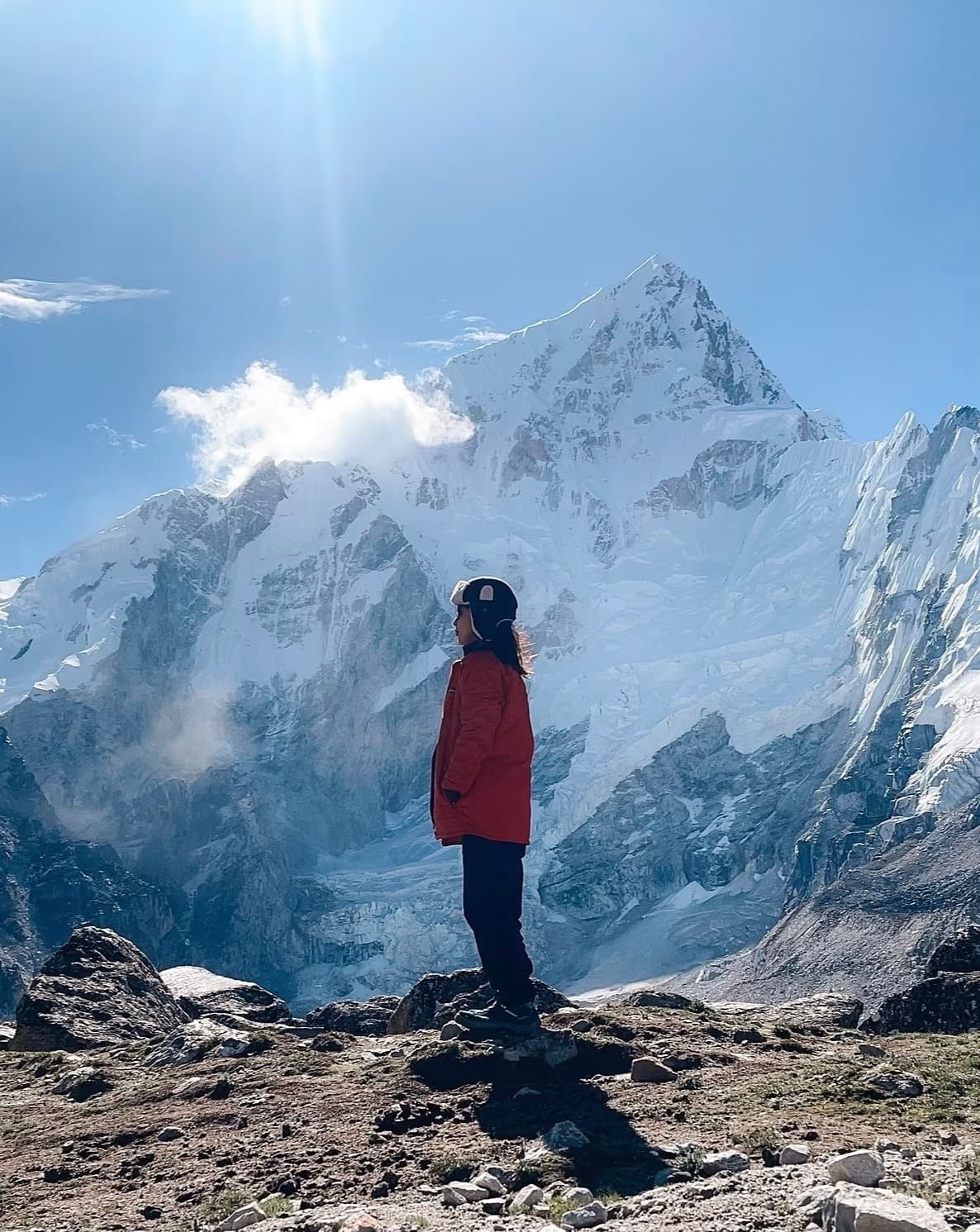 Maiya in a red jacket standing on rocky ground in front of a snow-covered mountain peak