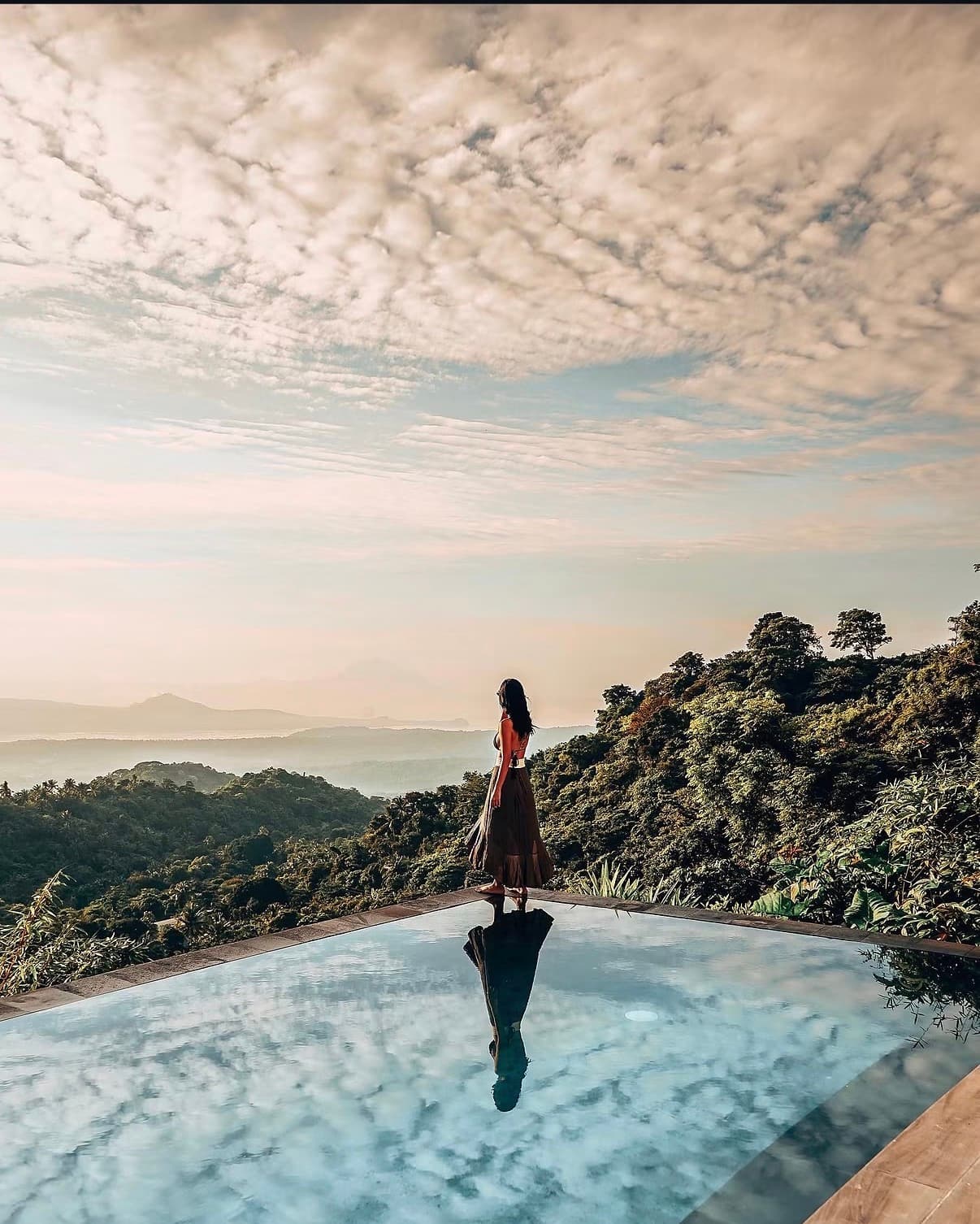 A woman standing at the edge of an infinity pool, looking out over forested hills under a cloudy sky